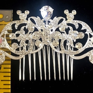 K&ouml;nnte beinhalten: Silberner Haarkamm mit Strassstein-Design mit zwei Mickey-Mouse-Ohren und einem gro&szlig;en, tropfenf&ouml;rmigen Strassstein in der Mitte. Der Kamm ist etwa 5 cm breit.