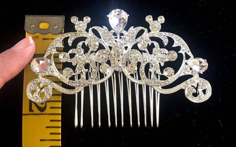 K&ouml;nnte beinhalten: Silberner Haarkamm mit Strassstein-Design mit zwei Mickey-Mouse-Ohren und einem gro&szlig;en, tropfenf&ouml;rmigen Strassstein in der Mitte. Der Kamm ist etwa 5 cm breit.