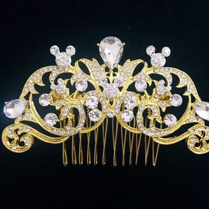 K&ouml;nnte beinhalten: Goldener Haarkamm mit einem dekorativen Design mit einer Krone, Schn&ouml;rkeln und Mickey-Maus-Silhouetten. Der Kamm ist mit klaren Strasssteinen verziert.