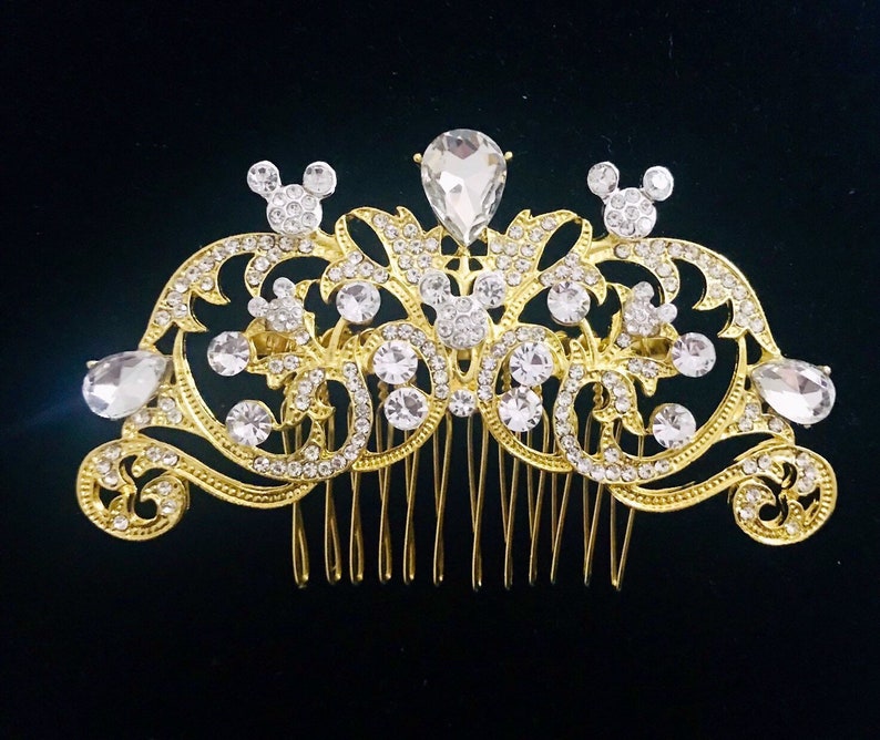 K&ouml;nnte beinhalten: Goldener Haarkamm mit einem dekorativen Design mit einer Krone, Schn&ouml;rkeln und Mickey-Maus-Silhouetten. Der Kamm ist mit klaren Strasssteinen verziert.