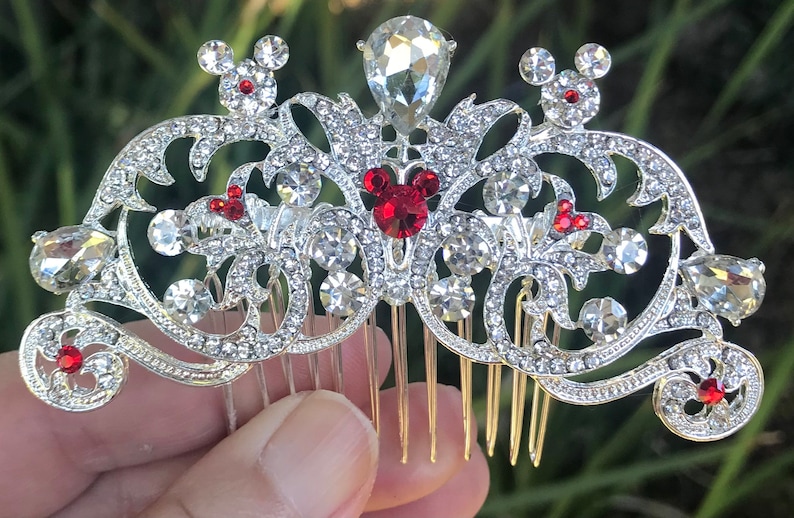 K&ouml;nnte beinhalten: Silberner Haarkamm mit einem dekorativen Design mit roten und klaren Strasssteinen. Das Design beinhaltet eine Mickey Mouse-Silhouette mit roten Ohren.