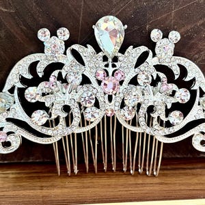 K&ouml;nnte beinhalten: Ein silberner Haarkamm mit einem dekorativen Design aus rosa und klaren Strasssteinen. Das Design beinhaltet einen gro&szlig;en zentralen Strassstein und zwei kleinere Strasssteine in Form von Mickey-Maus-K&ouml;pfen.