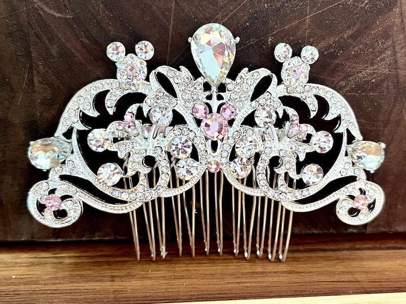 K&ouml;nnte beinhalten: Ein silberner Haarkamm mit einem dekorativen Design aus rosa und klaren Strasssteinen. Das Design beinhaltet einen gro&szlig;en zentralen Strassstein und zwei kleinere Strasssteine in Form von Mickey-Maus-K&ouml;pfen.