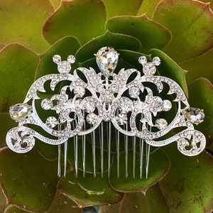 K&ouml;nnte beinhalten: Silberner Haarkamm mit einem dekorativen Design, das einen gro&szlig;en Kristall in der Mitte und kleinere Kristalle &uuml;berall enth&auml;lt. Das Design umfasst zwei Mickey-Maus-Ohren auf beiden Seiten des Kristalls.