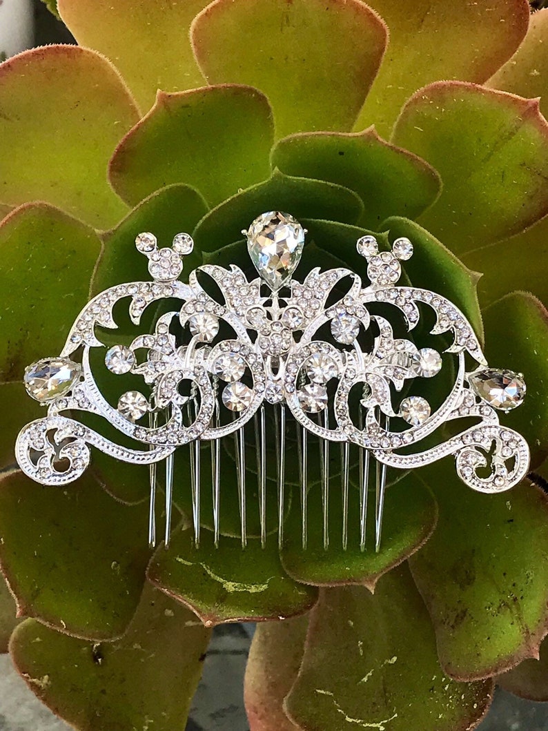 K&ouml;nnte beinhalten: Silberner Haarkamm mit einem dekorativen Design, das einen gro&szlig;en Kristall in der Mitte und kleinere Kristalle &uuml;berall enth&auml;lt. Das Design umfasst zwei Mickey-Maus-Ohren auf beiden Seiten des Kristalls.