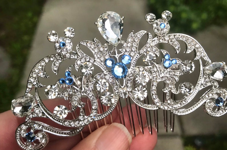 K&ouml;nnte beinhalten: Silberner Haarkamm mit floralem Design und blauen und klaren Strasssteinen. Der Kamm zeigt drei Mickey-Maus-Silhouetten.