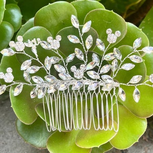 May include: A silver hair comb with a floral and leaf design. The comb features clear, sparkling crystals and is set against a backdrop of green succulent leaves. The comb's design includes small, round crystal clusters.