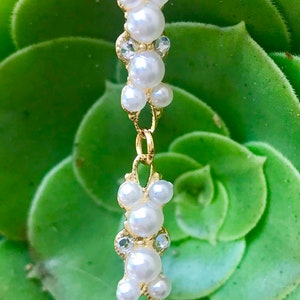 Puede incluir: Un brazalete de cadena dorada con racimos de perlas blancas y diamantes de imitaci&oacute;n transparentes. El brazalete est&aacute; sobre una planta suculenta verde.