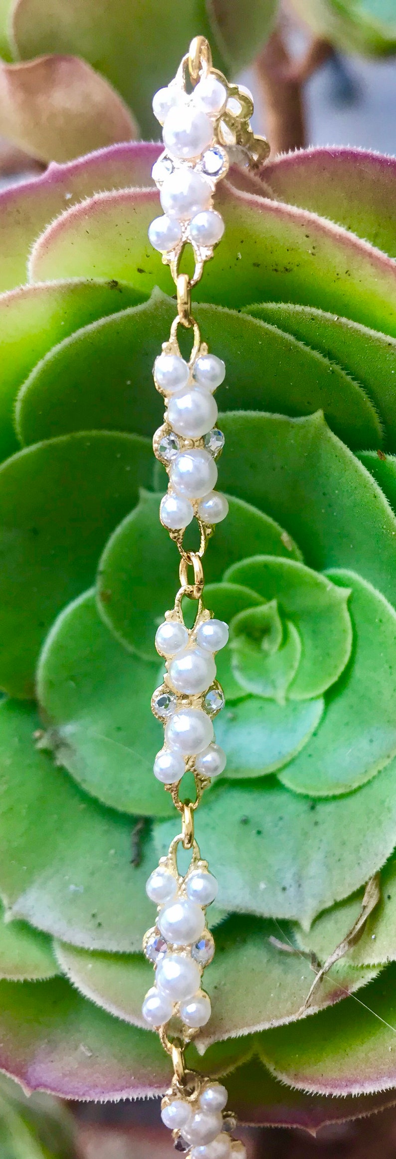 Puede incluir: Un brazalete de cadena dorada con racimos de perlas blancas y diamantes de imitaci&oacute;n transparentes. El brazalete est&aacute; sobre una planta suculenta verde.