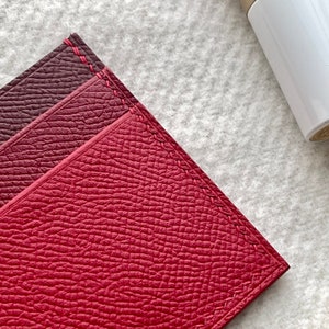 May include: Close-up of a red leather wallet with a burgundy leather flap. The wallet has a stitched edge and a textured surface.