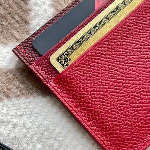 May include: A red leather wallet with a gold card inside. The wallet has a stitched edge and a textured surface.
