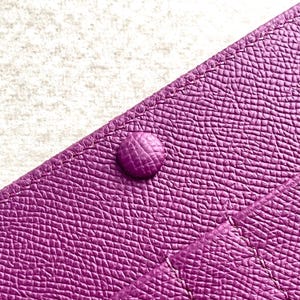 Puede incluir: Primer plano de una cartera de cuero p&uacute;rpura con un solo cierre a presi&oacute;n. La cartera tiene una superficie texturizada y un brillo sutil.