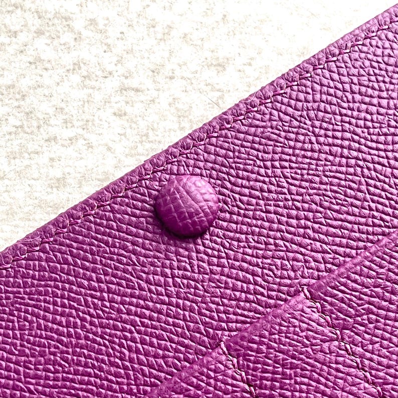 Puede incluir: Primer plano de una cartera de cuero p&uacute;rpura con un solo cierre a presi&oacute;n. La cartera tiene una superficie texturizada y un brillo sutil.