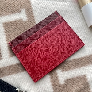 May include: A red leather card holder with a burgundy leather accent. The card holder has a stitched edge and a textured surface.