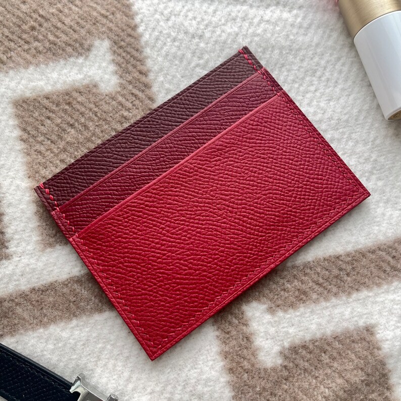 May include: A red leather card holder with a burgundy leather accent. The card holder has a stitched edge and a textured surface.