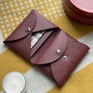 May include: A burgundy leather wallet with a snap closure, open to reveal a card and a portion of a banknote. The wallet is on a yellow and white patterned surface, with a candle and a red container in the background.