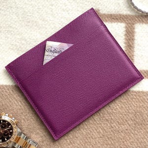 Puede incluir: Un porta tarjetas de cuero morado con un solo bolsillo. El porta tarjetas est&aacute; sobre una superficie con un patr&oacute;n beige y marr&oacute;n.