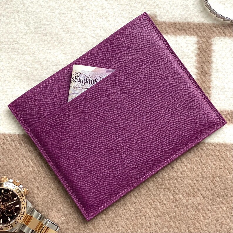 Puede incluir: Un porta tarjetas de cuero morado con un solo bolsillo. El porta tarjetas est&aacute; sobre una superficie con un patr&oacute;n beige y marr&oacute;n.