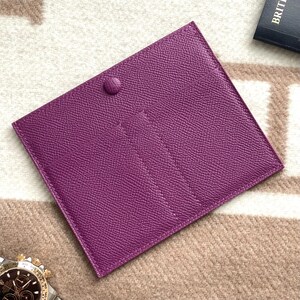 Puede incluir: Una cartera de cuero p&uacute;rpura con m&uacute;ltiples ranuras para tarjetas y un cierre a presi&oacute;n.