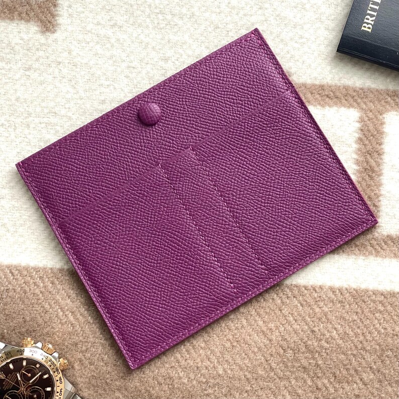 Puede incluir: Una cartera de cuero p&uacute;rpura con m&uacute;ltiples ranuras para tarjetas y un cierre a presi&oacute;n.