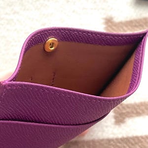 Puede incluir: Un tarjetero de cuero morado con cierre de presi&oacute;n dorado. El interior del tarjetero es de cuero marr&oacute;n.