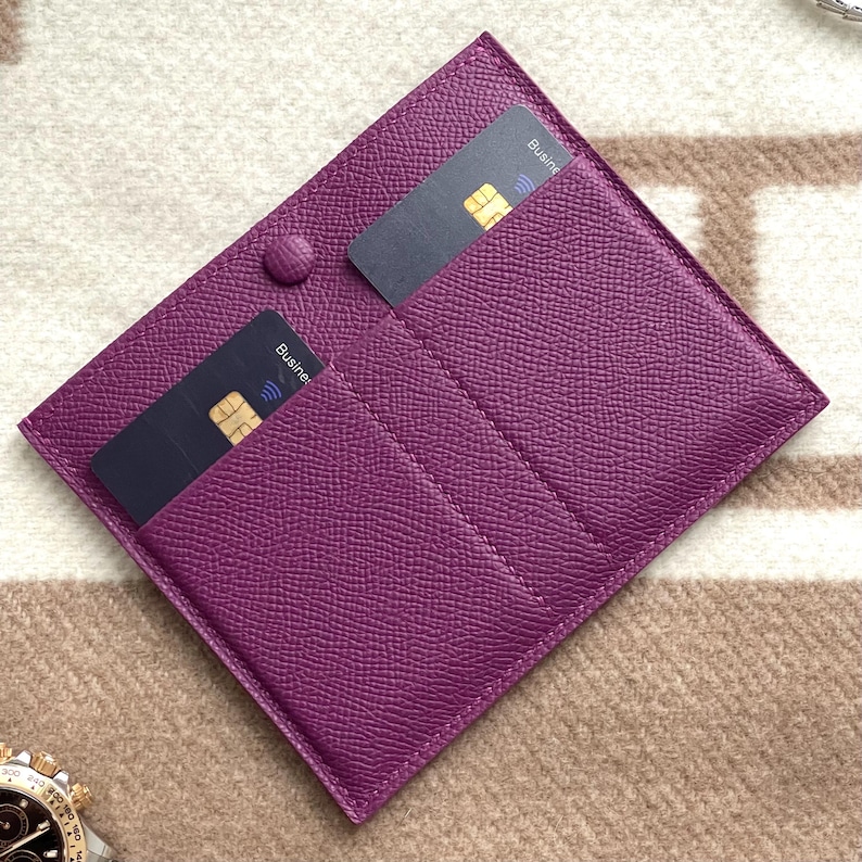 Puede incluir: Un porta tarjetas de cuero morado con dos tarjetas negras en su interior. Las tarjetas tienen el texto "Business" impreso en ellas.