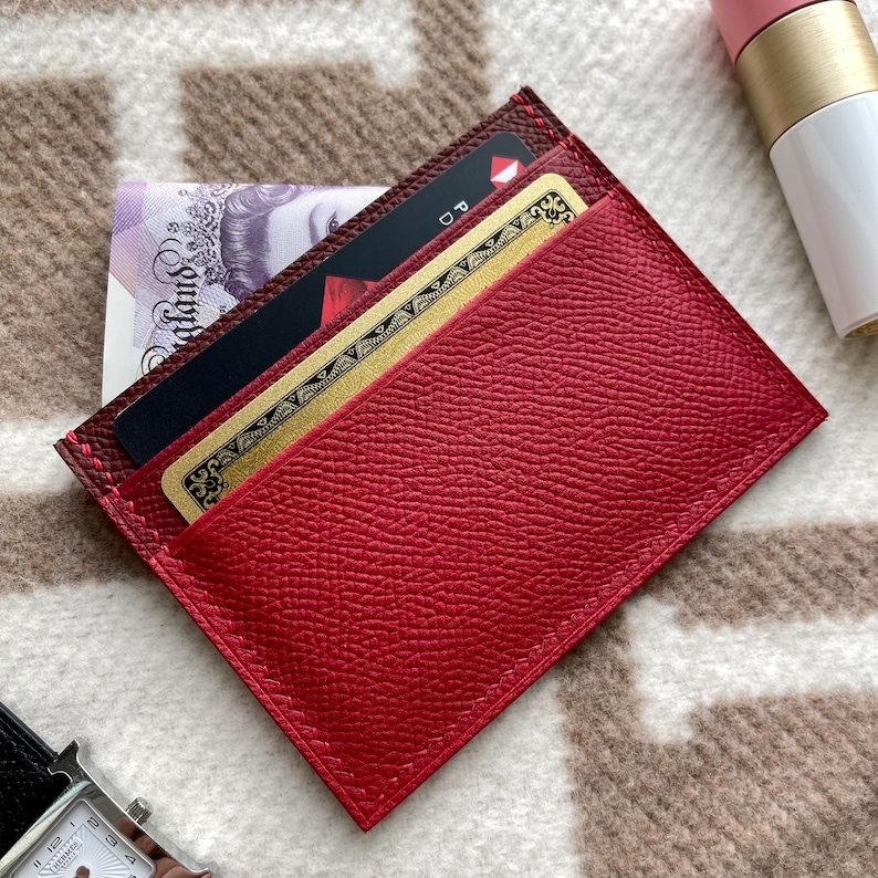 May include: A red leather card holder with two cards inside. The top card is a gold credit card with a black border and the bottom card is a black playing card with a red diamond.