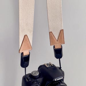 May include: A beige canvas camera strap with brown leather accents and black plastic clips. The strap is attached to a black Canon camera. The Canon logo is visible on the camera body.
