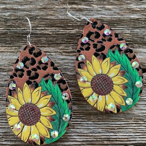 May include: A pair of dangle earrings with a brown and black leopard print background. Each earring features a yellow sunflower with green leaves and clear rhinestones.