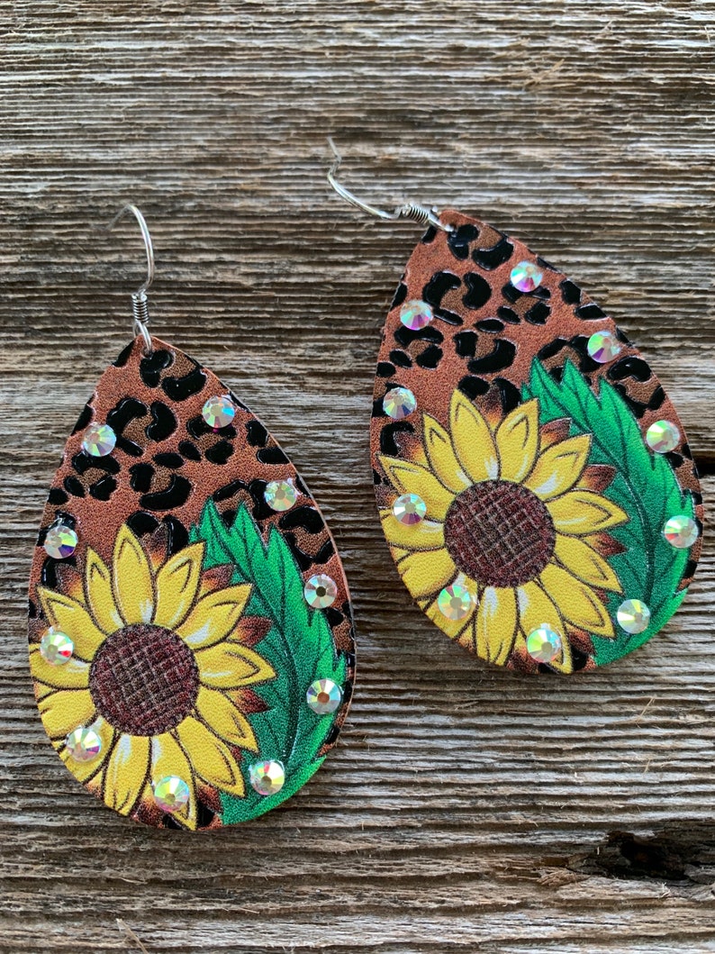 May include: A pair of dangle earrings with a brown and black leopard print background. Each earring features a yellow sunflower with green leaves and clear rhinestones.