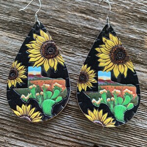 May include: A pair of dangle earrings with a black background and a Texas state outline. The earrings are decorated with yellow sunflowers and green cactus plants.