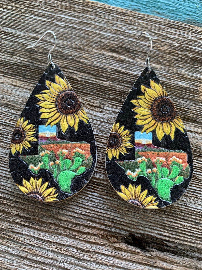 May include: A pair of dangle earrings with a black background and a Texas state outline. The earrings are decorated with yellow sunflowers and green cactus plants.