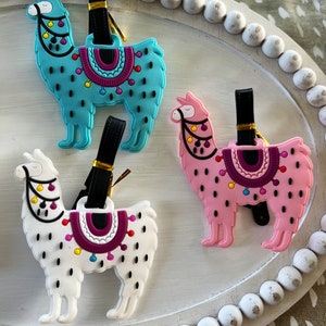 May include: Three colorful llama luggage tags with black straps. The llamas are turquoise, pink, and white with black spots and colorful accents.