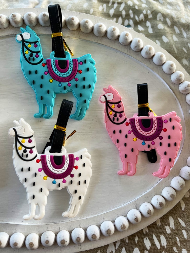 May include: Three colorful llama luggage tags with black straps. The llamas are turquoise, pink, and white with black spots and colorful accents.