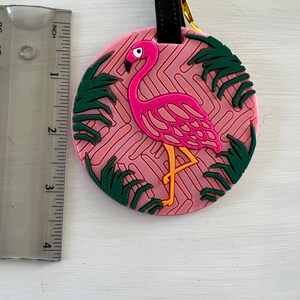Op de afbeelding: Roze flamingo bagagetag met groene palmbladeren op een roze en witte gepatroneerde achtergrond. De tag heeft een zwarte band met een gouden metalen lus.