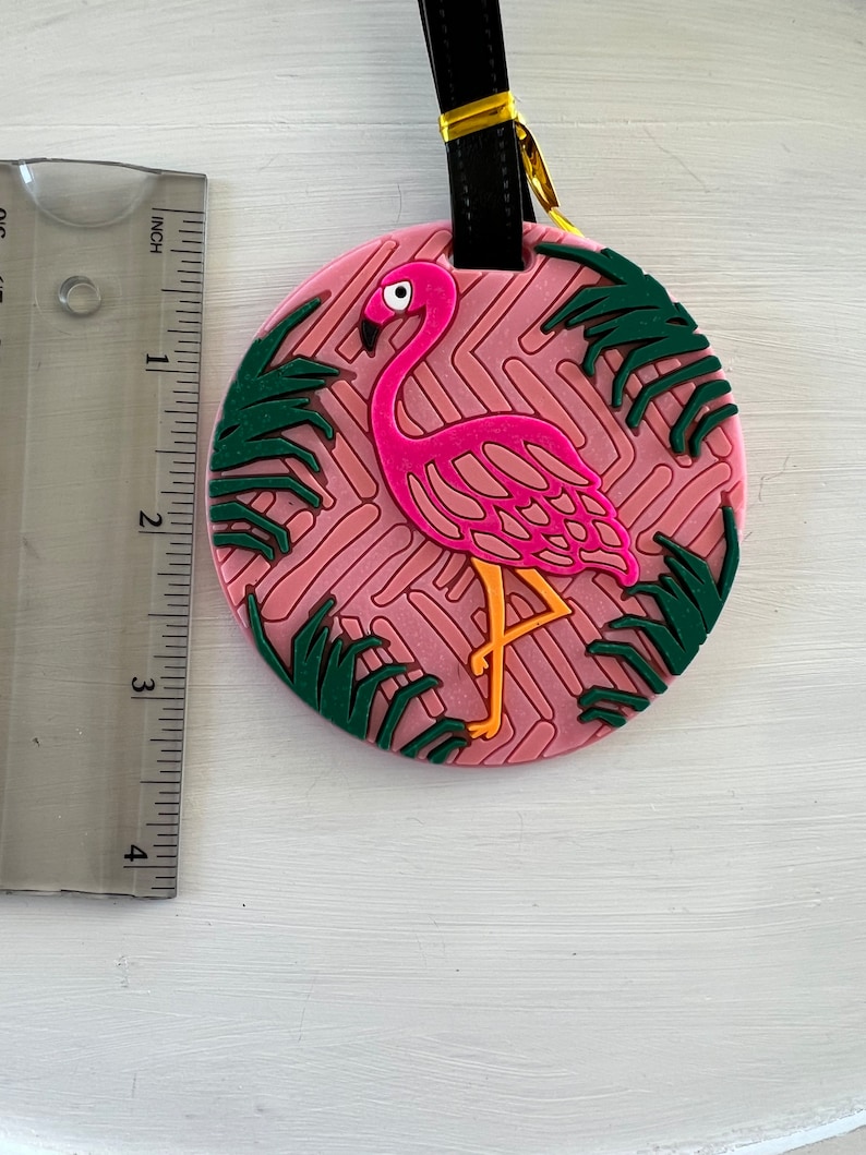 Op de afbeelding: Roze flamingo bagagetag met groene palmbladeren op een roze en witte gepatroneerde achtergrond. De tag heeft een zwarte band met een gouden metalen lus.