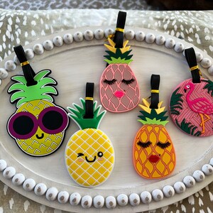 Op de afbeelding: Vijf kleurrijke ananas- en flamingo-bagagetags met zwarte bandjes. De ananas-tags hebben verschillende gezichtsuitdrukkingen, waaronder een zonnebril, wimpers en een knipoog. De flamingo-tag is roze met groene palmbladeren.