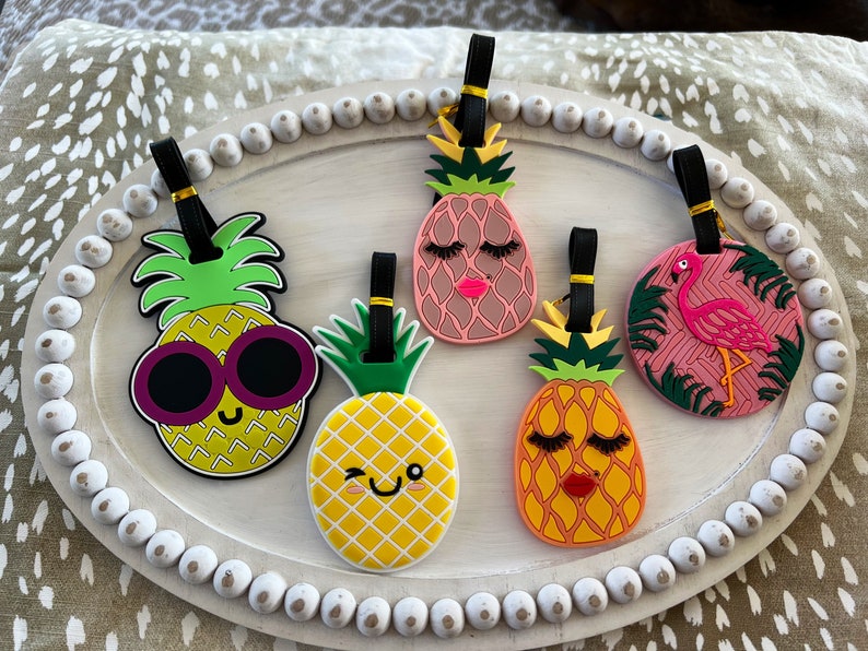 Op de afbeelding: Vijf kleurrijke ananas- en flamingo-bagagetags met zwarte bandjes. De ananas-tags hebben verschillende gezichtsuitdrukkingen, waaronder een zonnebril, wimpers en een knipoog. De flamingo-tag is roze met groene palmbladeren.