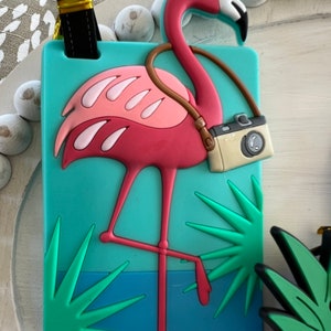 K&ouml;nnte beinhalten: Rosafarbener Flamingo-Gep&auml;ckanh&auml;nger mit Kameragurt und gr&uuml;nen Palmenbl&auml;ttern auf t&uuml;rkisfarbenem Hintergrund.