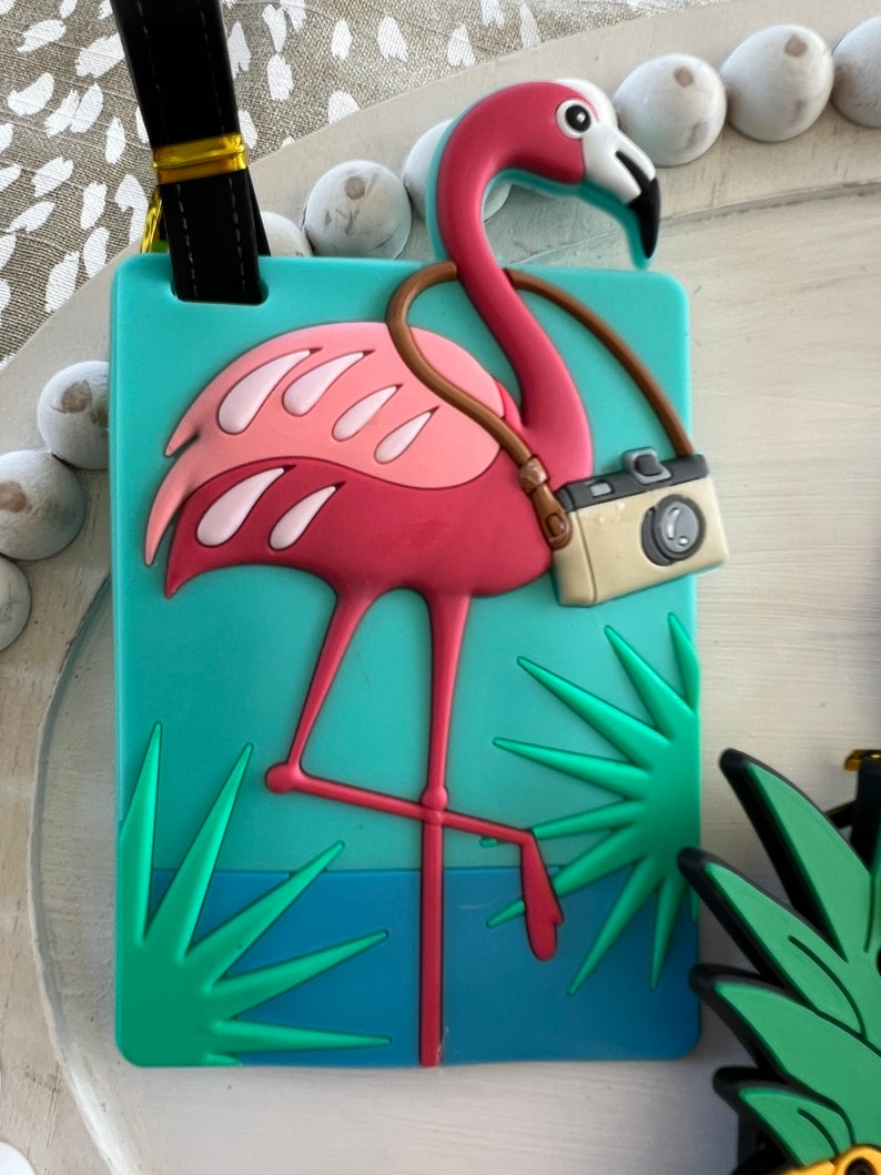 K&ouml;nnte beinhalten: Rosafarbener Flamingo-Gep&auml;ckanh&auml;nger mit Kameragurt und gr&uuml;nen Palmenbl&auml;ttern auf t&uuml;rkisfarbenem Hintergrund.