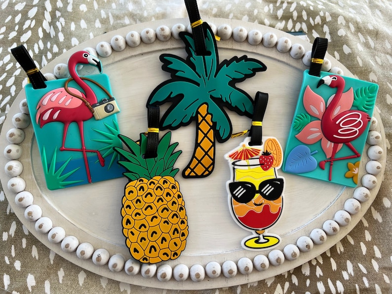 K&ouml;nnte beinhalten: Vier farbenfrohe Gep&auml;ckanh&auml;nger mit tropischen Motiven. Ein rosa Flamingo mit einer Kamera, eine Ananas, eine Palme und ein Cocktail mit Sonnenbrille.