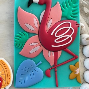 K&ouml;nnte beinhalten: Ein t&uuml;rkisfarbener Gep&auml;ckanh&auml;nger mit einem rosa Flamingo, gr&uuml;nen Bl&auml;ttern und einem blauen Blatt. Der Flamingo hat wei&szlig;e Akzente und steht auf einem Bein.