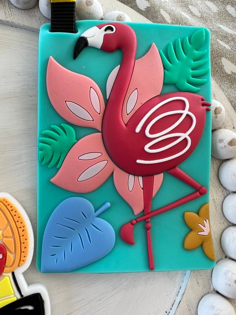 K&ouml;nnte beinhalten: Ein t&uuml;rkisfarbener Gep&auml;ckanh&auml;nger mit einem rosa Flamingo, gr&uuml;nen Bl&auml;ttern und einem blauen Blatt. Der Flamingo hat wei&szlig;e Akzente und steht auf einem Bein.