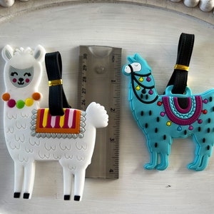 May include: Two colorful llama luggage tags. The white llama has a pink, yellow, and orange saddle and a colorful beaded necklace. The blue llama has a purple, pink, and yellow saddle with black polka dots. Both llamas have black eyes and a black nose.