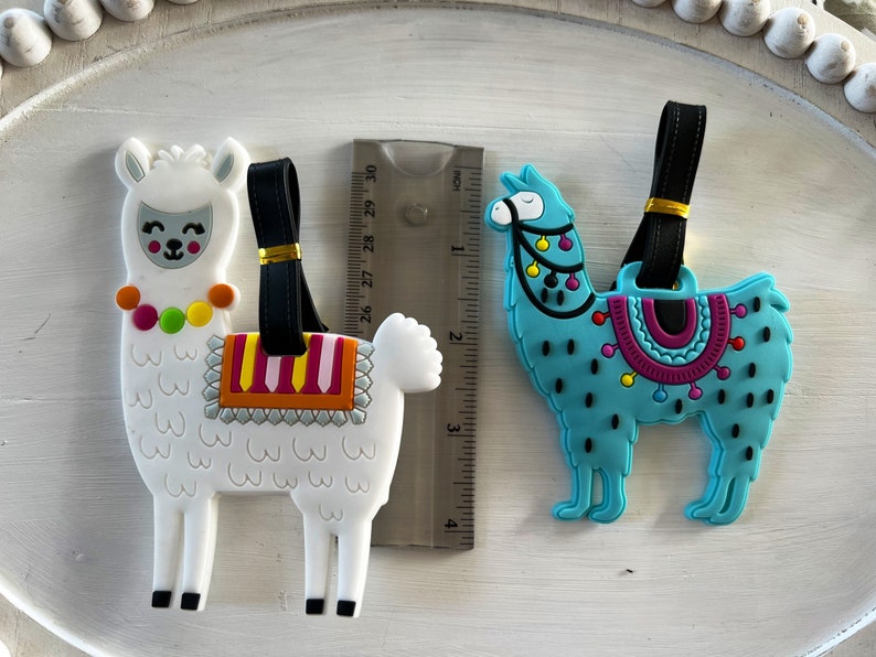 May include: Two colorful llama luggage tags. The white llama has a pink, yellow, and orange saddle and a colorful beaded necklace. The blue llama has a purple, pink, and yellow saddle with black polka dots. Both llamas have black eyes and a black nose.