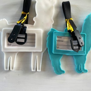 May include: Two llama-shaped luggage tags with black straps and gold accents. The tags have blank spaces for writing names, addresses, and phone numbers. One tag is white and the other is blue.