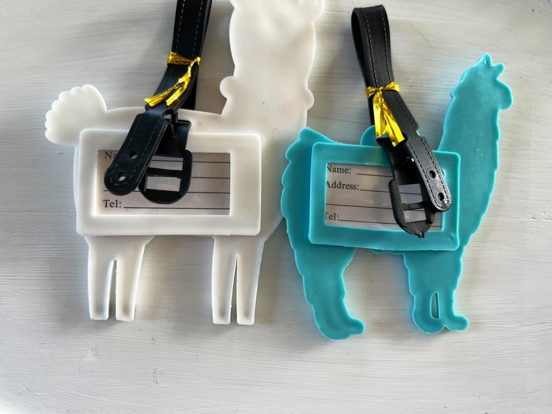 May include: Two llama-shaped luggage tags with black straps and gold accents. The tags have blank spaces for writing names, addresses, and phone numbers. One tag is white and the other is blue.