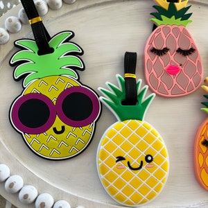 Op de afbeelding: Vier kleurrijke ananasvormige bagagelabeltjes met zwarte bandjes en gouden accenten. De ananas hebben verschillende gezichtsuitdrukkingen en details, waaronder een zonnebril, wimpers en een knipoog.
