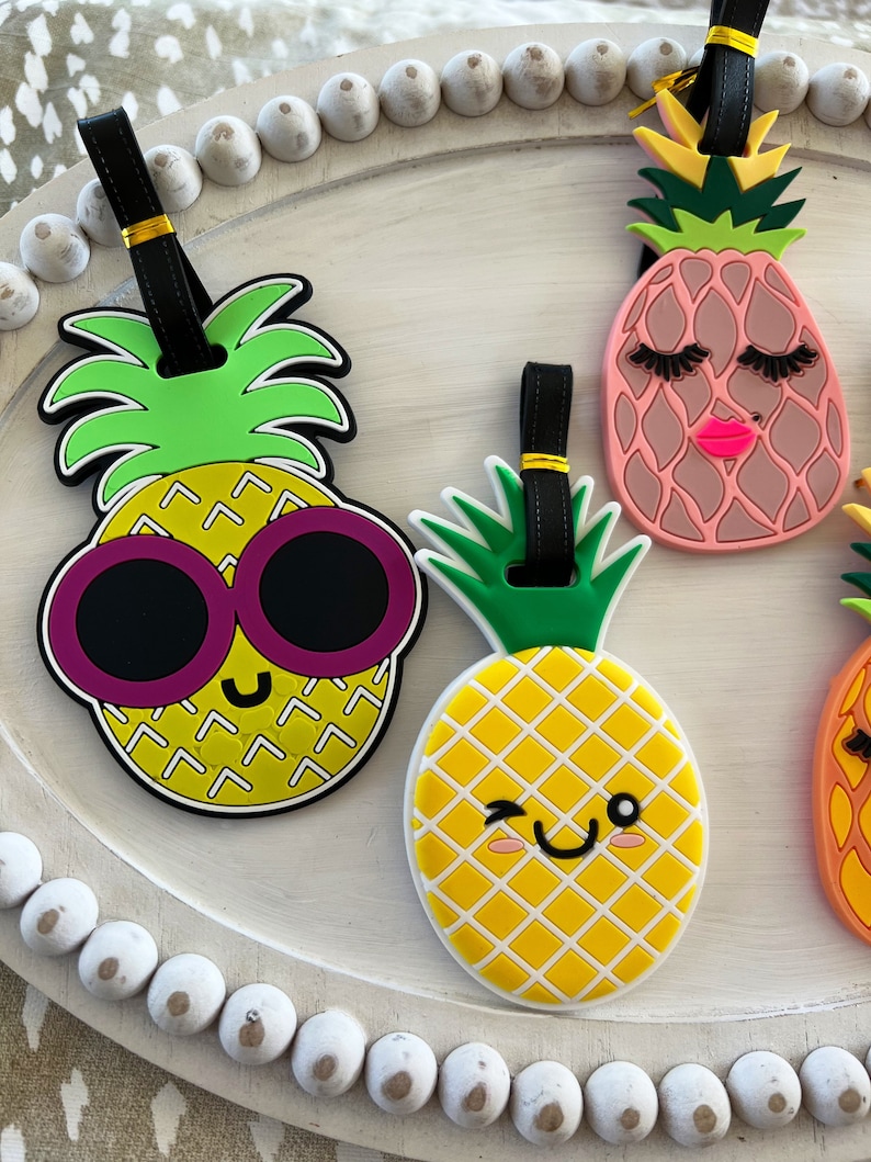 Op de afbeelding: Vier kleurrijke ananasvormige bagagelabeltjes met zwarte bandjes en gouden accenten. De ananas hebben verschillende gezichtsuitdrukkingen en details, waaronder een zonnebril, wimpers en een knipoog.