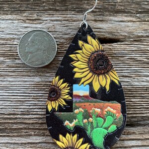 May include: A pair of dangle earrings featuring a black leather teardrop shape with a Texas outline and cactus design. The earrings are decorated with a sunflower pattern.